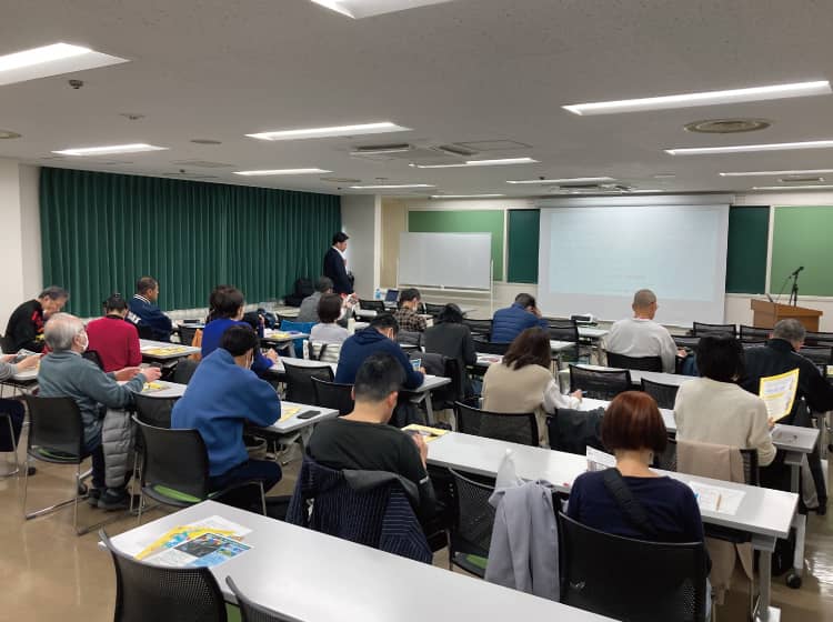 Adult students seated at long desks in a classroom, a presenter stands near whiteboards and a projection screen at the front.